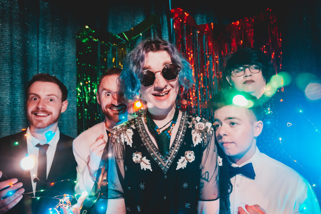 A photo of the members of Strange New Places, dressed in formal wear, shrouded in fairy lights and in front of a glittery backdrop. Ash wears mirrored sunglasses and everyone looks unhinged.